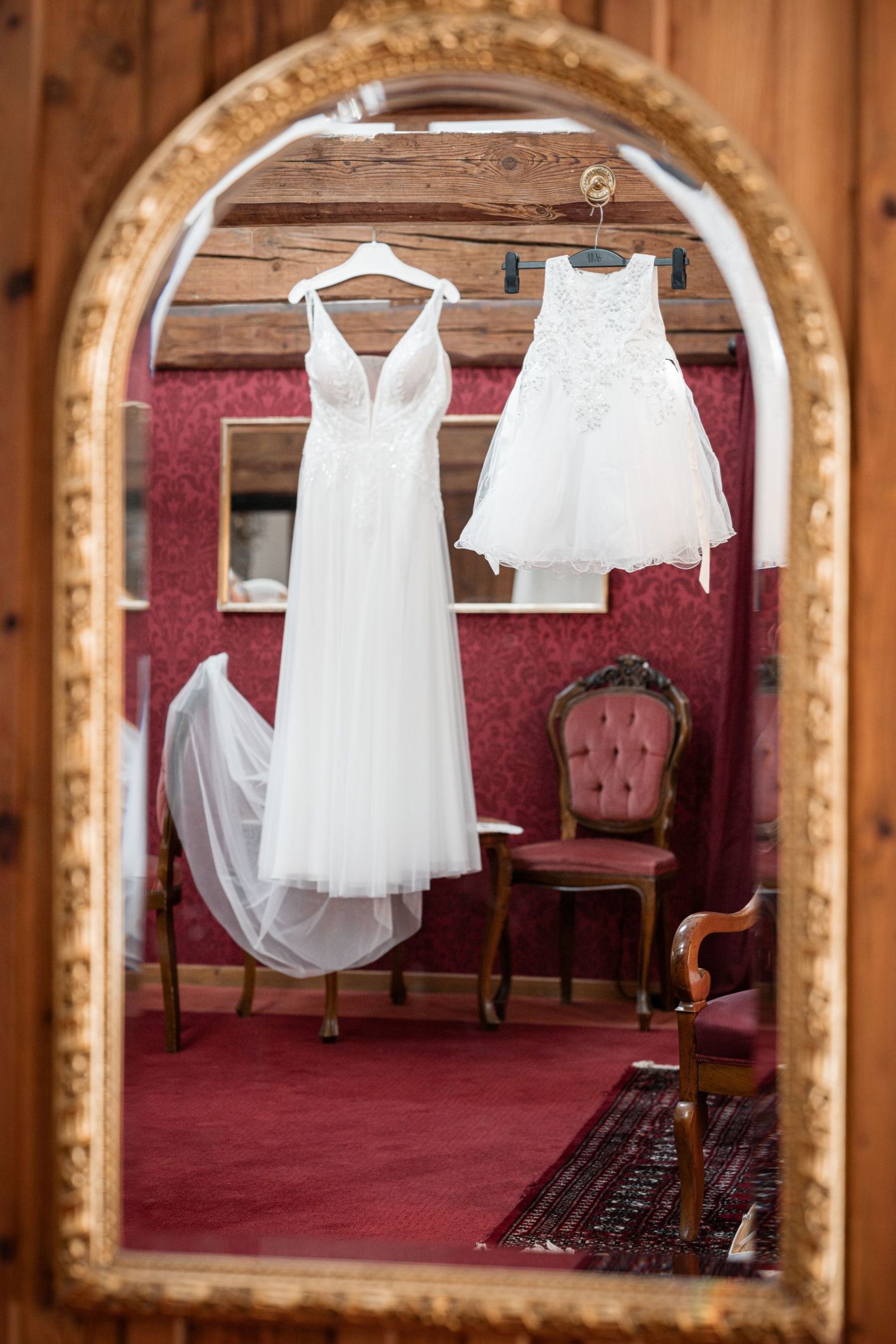 Detailaufnahme des Brautkleids und des kleinen weißen Kleides der Tochter, kunstvoll durch den Spiegel im roten Ankleidezimmer des Vedahof aufgenommen.