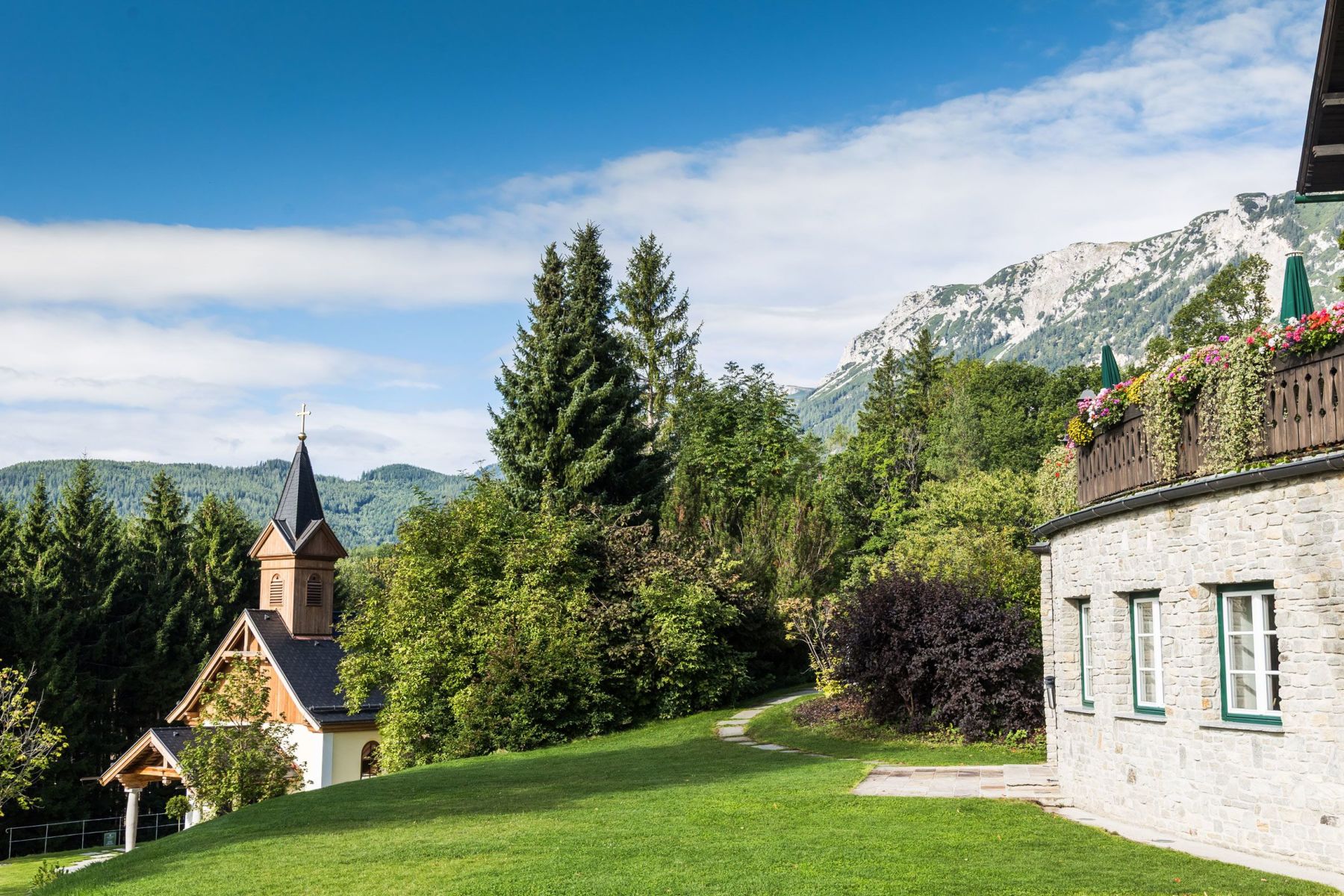 Blick auf das Haus und die Kapelle am Knappenhof mit der sommerlichen Landschaft und dem Rax-Gebirge im Hintergrund.